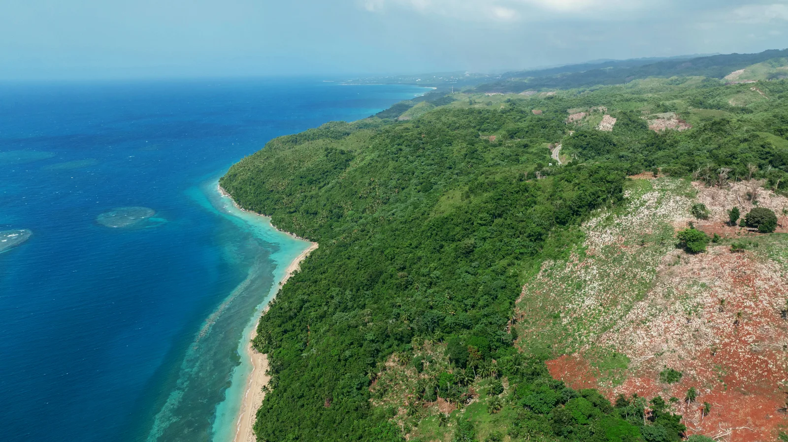 Vista aérea del terreno: lomas boscosas y una ladera desbrozada de tierra roja que baja hasta una playa y un arrecife